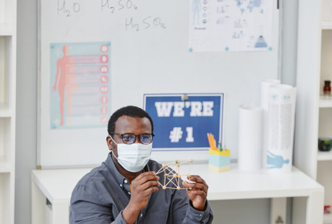 Vertical portrait of African American teacher wearing mask and holding molecule model during chemistry class in school