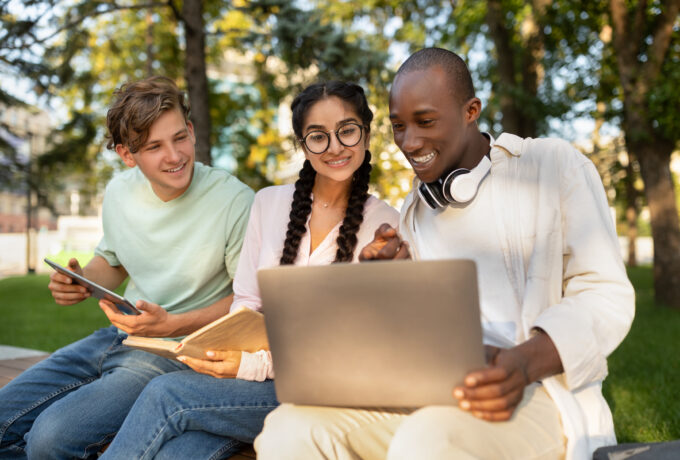 Excited international students studying in park, enjoying the results of their job, looking at laptop screen and smiling, sitting together on bench in campus