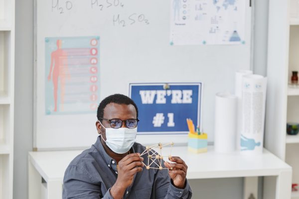 Vertical portrait of African American teacher wearing mask and holding molecule model during chemistry class in school